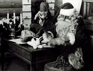 santa sits at a desk writing his list while an elf answers a phone next to him
