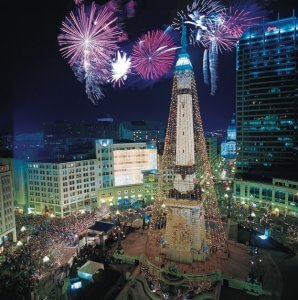 Tall white monument decorated by Christmas lights in Christmas tree shape in middle of town surrounded by buildings and dense crowd with red fireworks in background